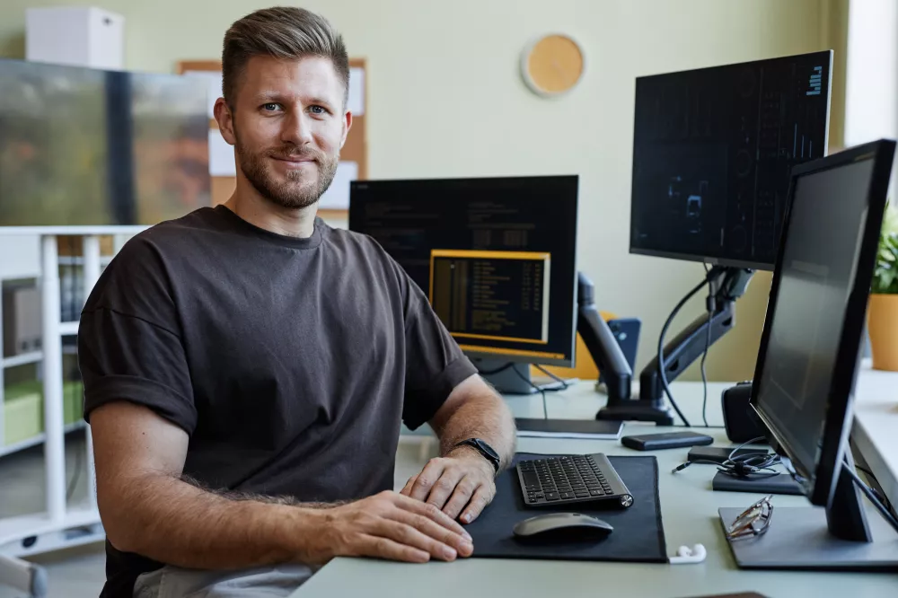 portrait-smiling-software-developer-looking-camera-while-sitting-desk-with-computers