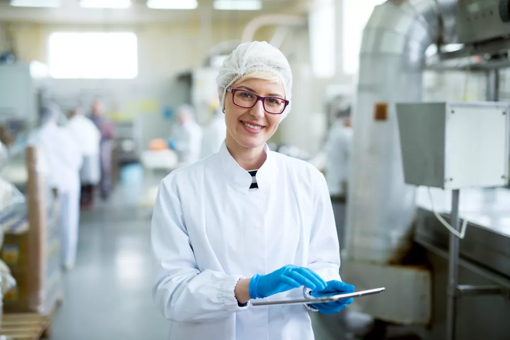 young-joyful-beautiful-female-worker-sterile-cloths-holding-tablet-smiling-camera-near-factory-production-line